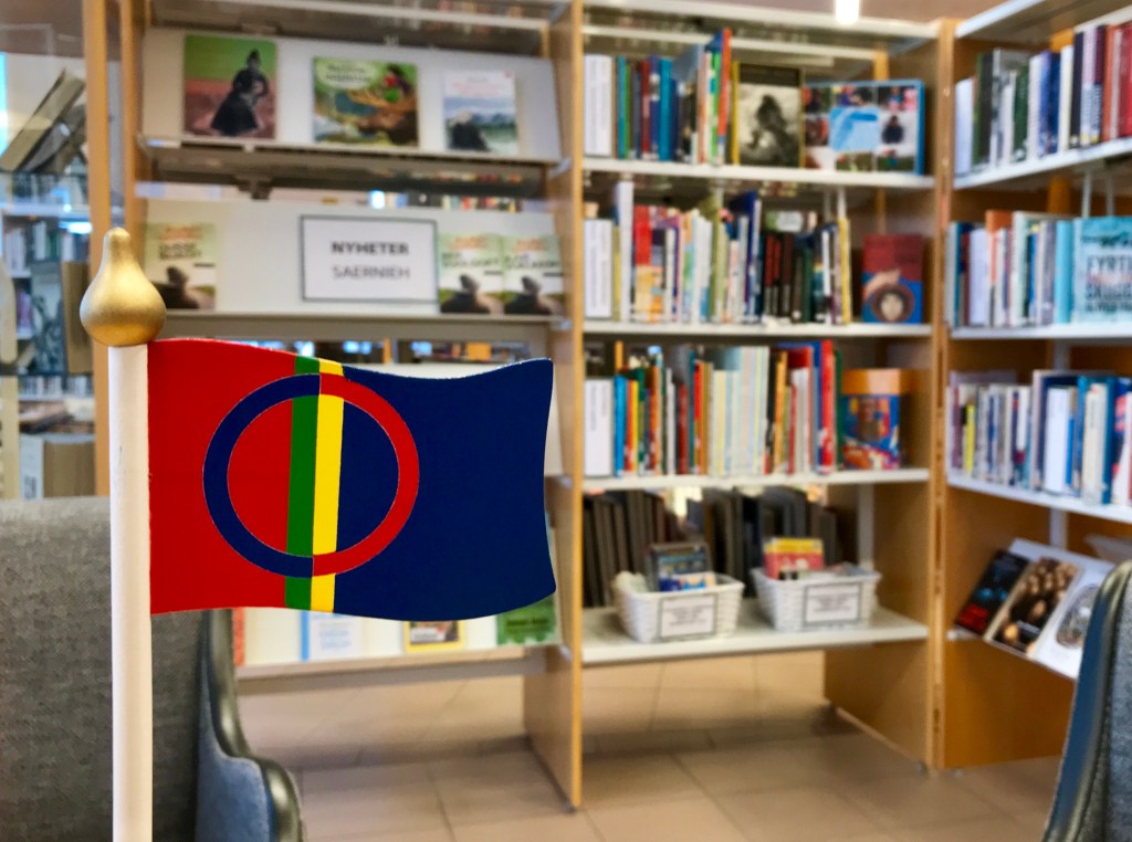 Biblioteksrum, böcker i bakgrunden, en liten samisk flagga i trä i förgrunden. 