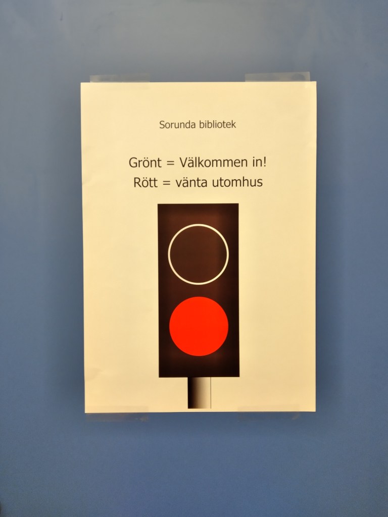 Ett trafikljus lyser rött. En text står bredvid: Sorunda bibliotek Grönt = välkommen in, Rött = Vänta utomhus.